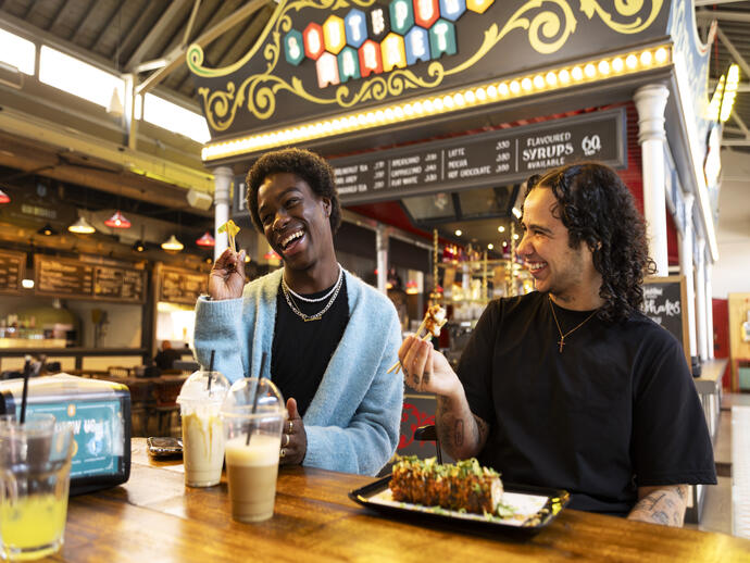 Two friends laugh while eating and drinking inside a food market