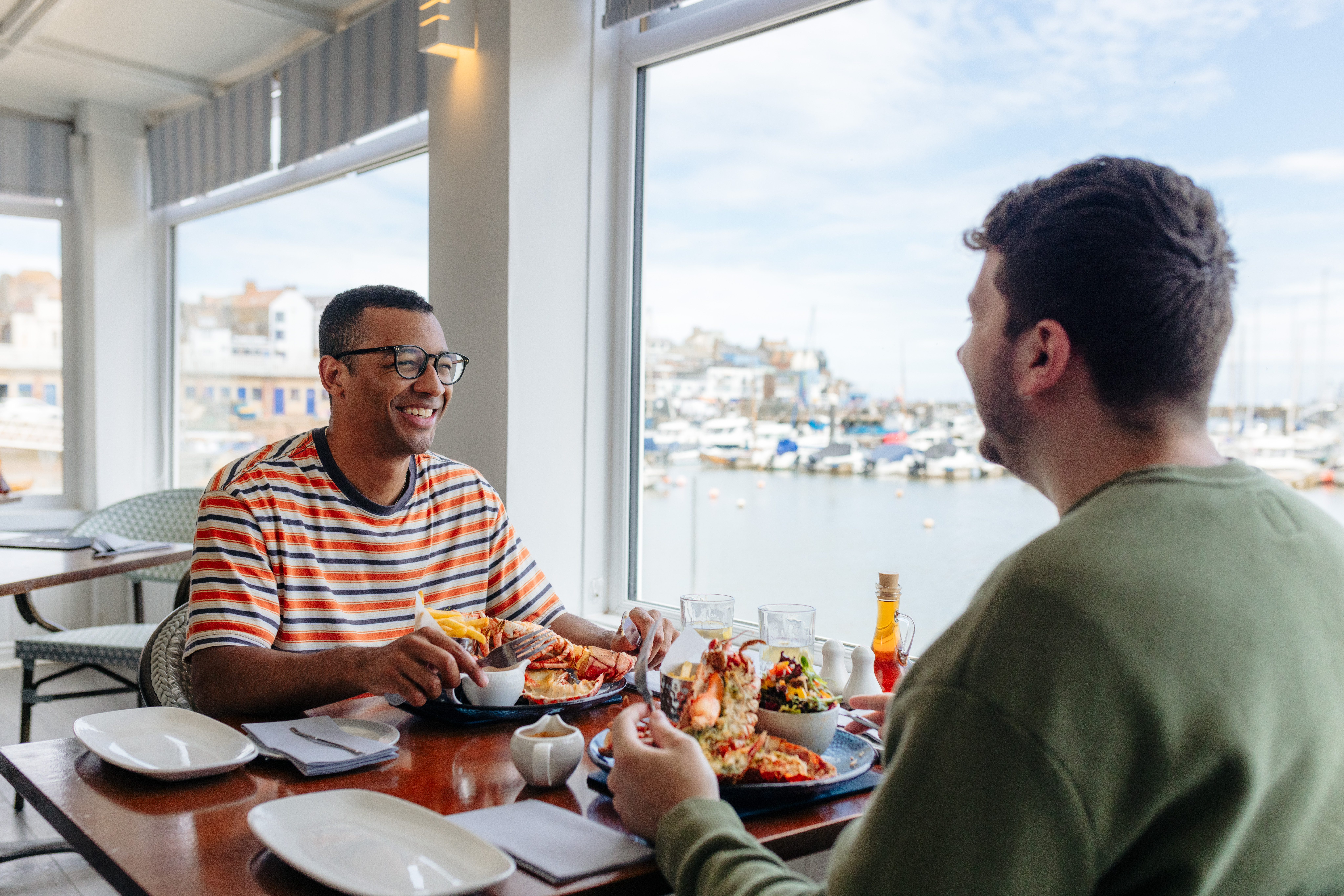 Two men eat lobster in a restaurant with a harbour view
