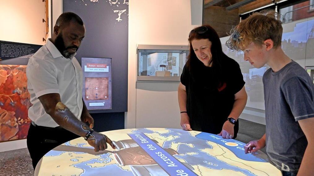 Three people interact with a large touchscreen display map in a modern museum, engaging with educational content about the Roman Empire.