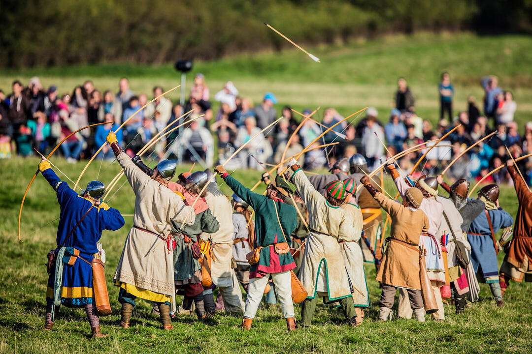 People in historical costumes reenact archery, drawing bows in a field with an audience in the background.