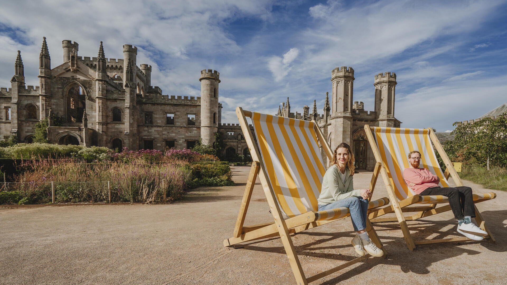 Deux personnes assises sur d’énormes chaises longues rayées jaune et blanc devant un grand château historique avec tours et jardins sous un ciel bleu.