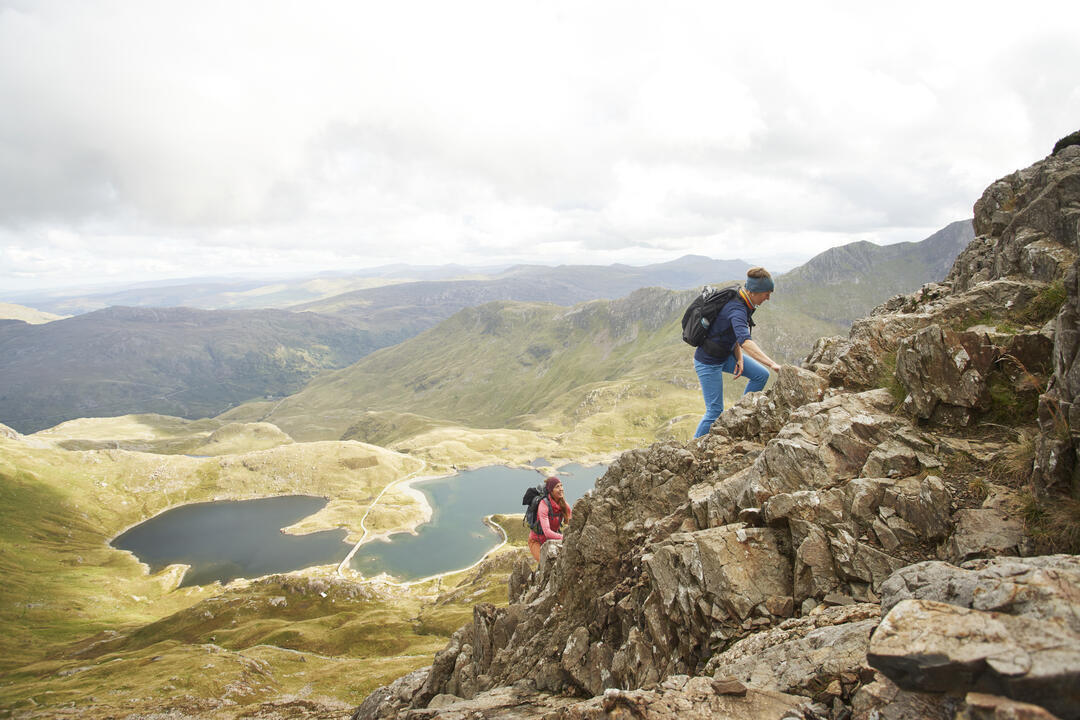 Two females hiking together up a large mountain range, with a lake below