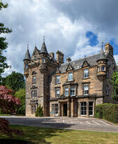 Historic stone castle-like mansion with turrets, surrounded by trees and a manicured lawn under a partly cloudy sky.