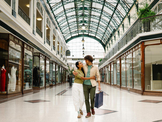 A couple walks inside a historic shopping arcade with a large glass domed roof, surrounded by elegant shopfronts and mannequins on display.
