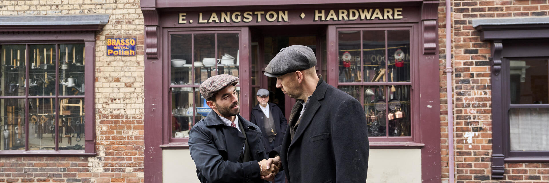 Two men in vintage attire shake hands outside a brick hardware store named E. Langston. Historical street scene with older shopfronts.