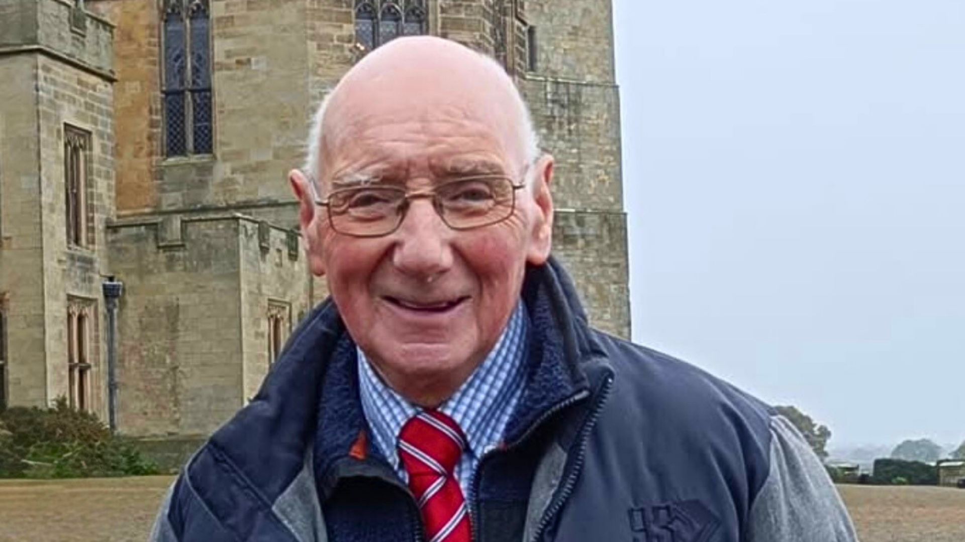 Smiling older man with glasses standing outside a historic stone building on a cloudy day.