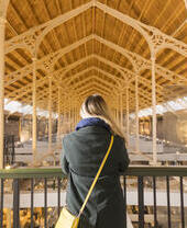 Person with a yellow bag stands on a balcony, overlooking a spacious hall with a high wooden ceiling and glass windows.