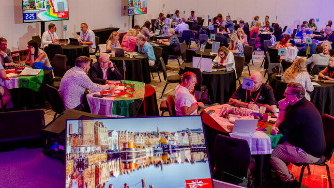 A conference room with round tables, people networking, laptops open, and a screen showing an image of Edinburgh, Scotland. Professional event atmosphere.
