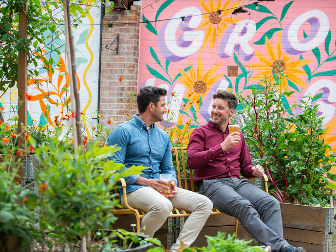 Two men sat on a bench in a garden with a drink