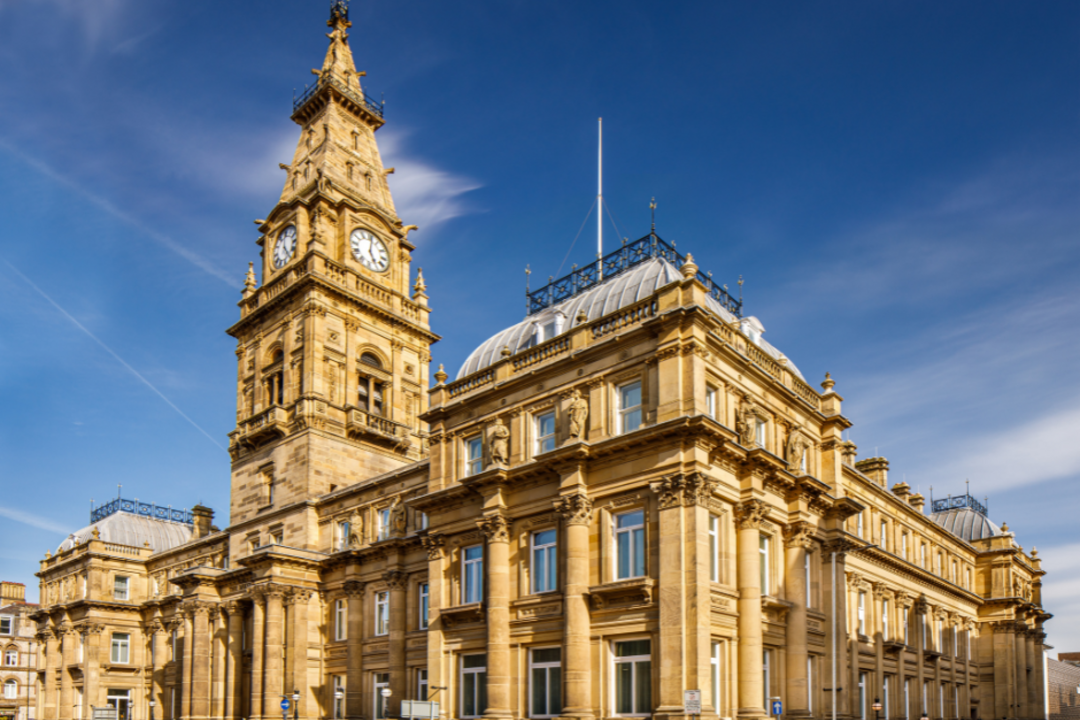 Historic clock tower and ornate stone building with domed roof under blue sky, cars parked along the street.