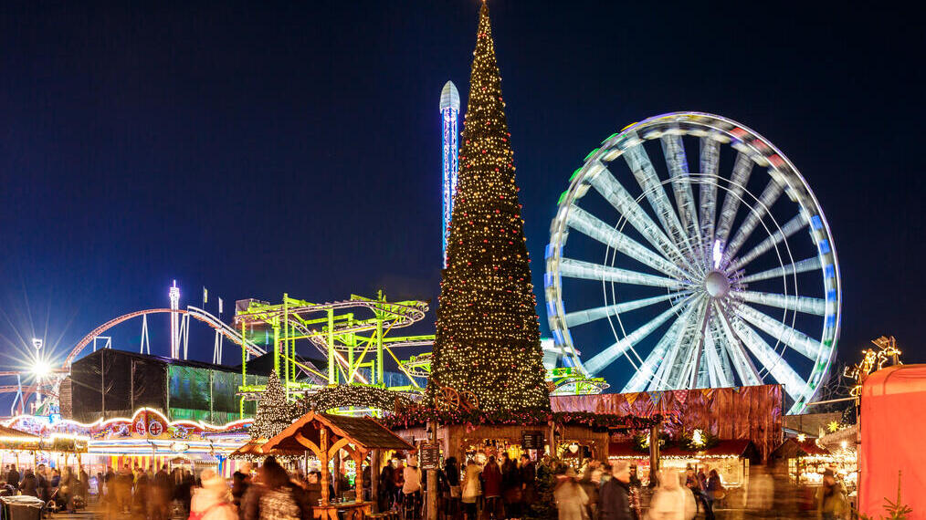 Ein Weihnachtsmarkt bei Nacht mit großem geschmücktem Weihnachtsbaum, Riesenrad, Achterbahn, Lichtern und Menschenmengen.