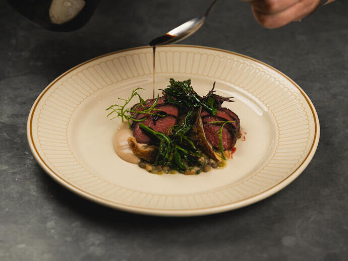 Plated gourmet dish being garnished with sauce, featuring sliced meat, greens, and a creamy puree on a patterned plate.