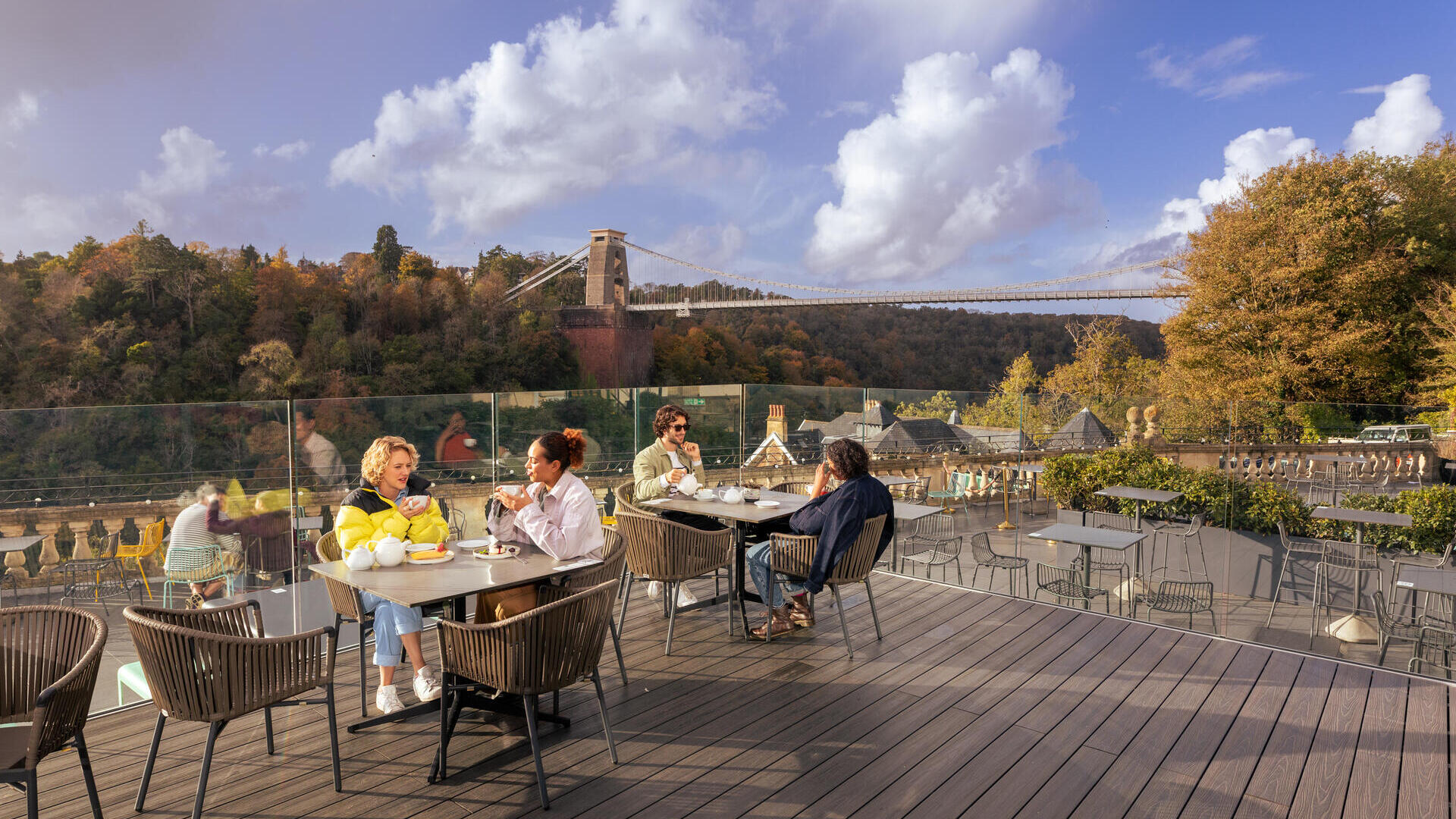 Des amis dégustant du thé et des gâteaux sur une terrasse sur le toit, avec vue sur une rivière et un pont suspendu.