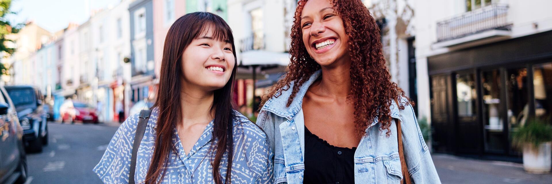 Zwei junge Frauen, die Arm in Arm lächelnd eine Straße entlanggehen