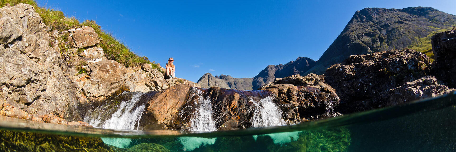 Clear stream with visible underwater rocks, small waterfall, rocky landscape and mountains under a blue sky. Person sits on rocks above the water.