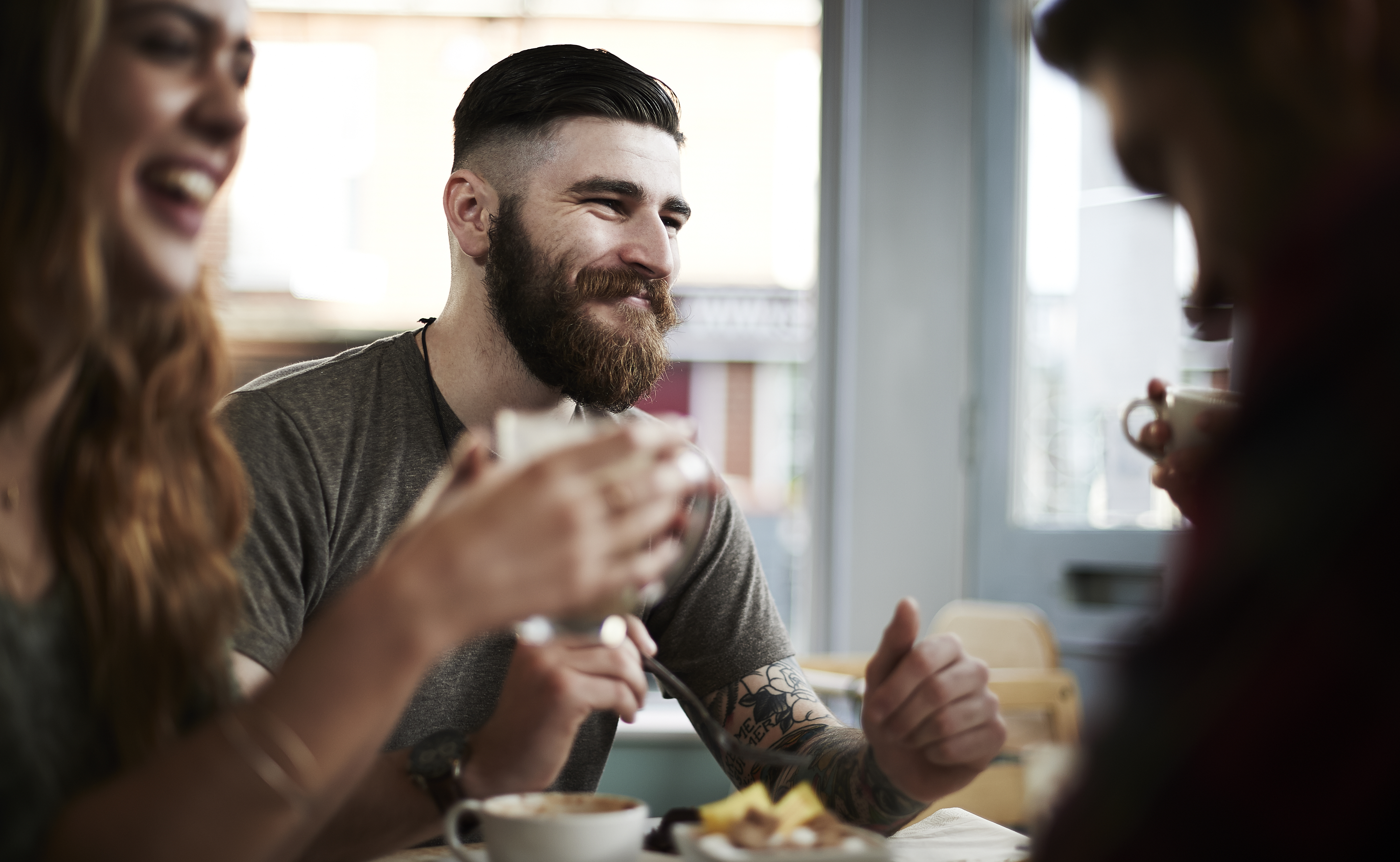 Young bearded male with tattoos socializing with group of friends in cafe.