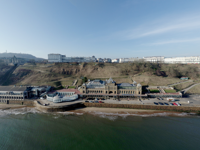 Seafront view of historic grand buildings on the coast, sandy beach, cliffs, greenery and clear blue sky.