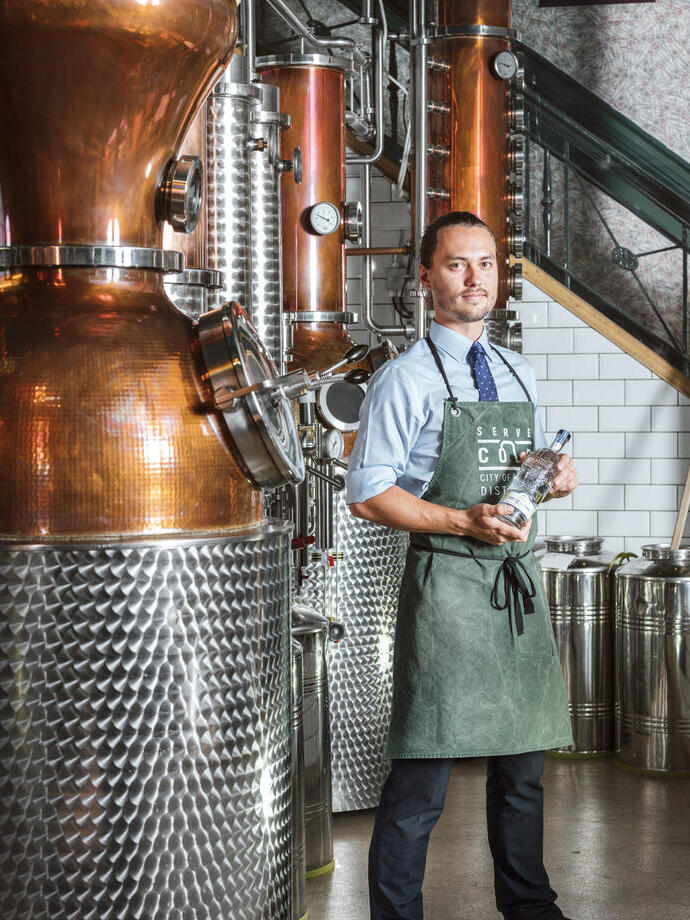 Man wearing green apron standing next to a copper still in a gin distillery