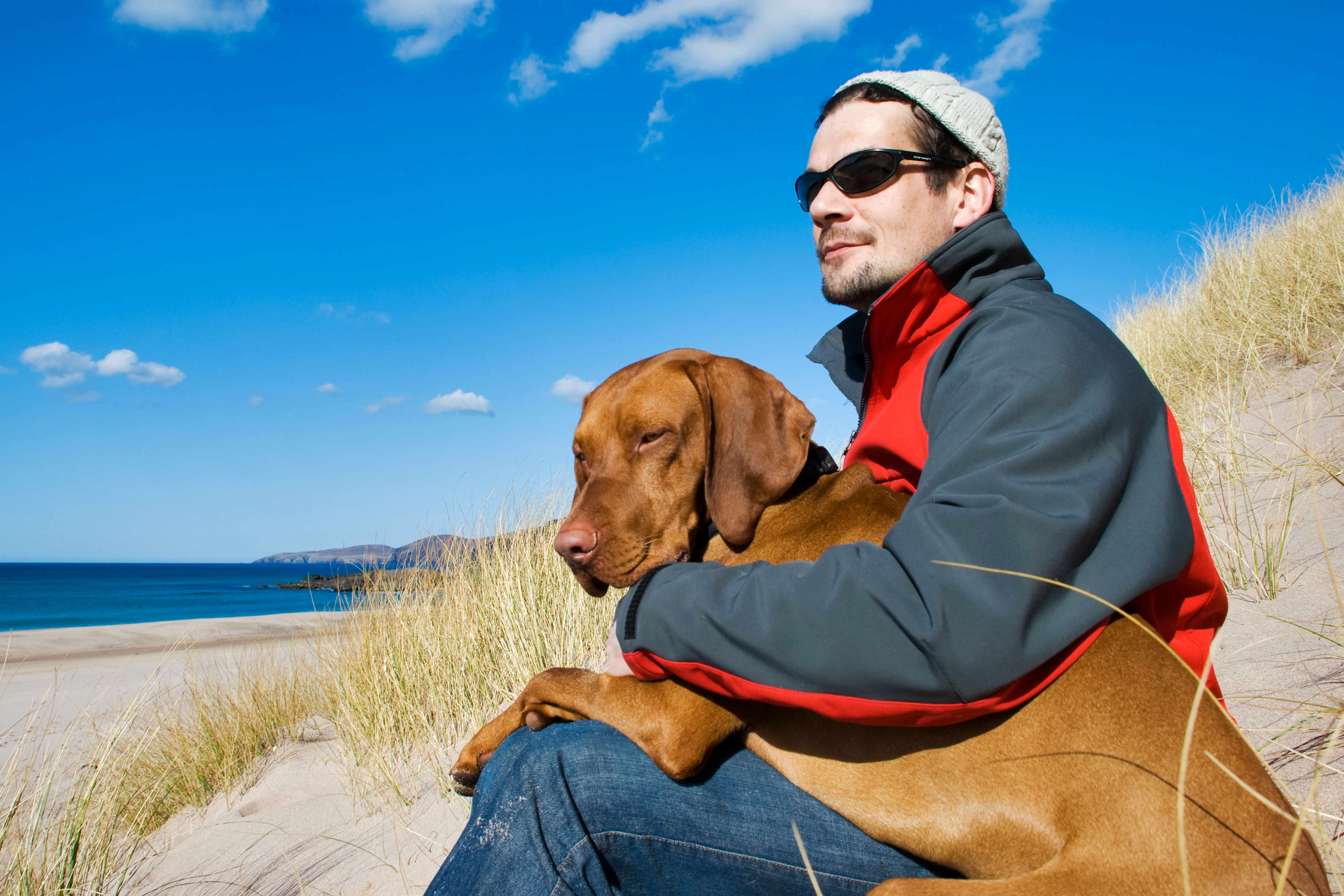 Mann sitzt mit einem ungarischen Vizsla-Hund auf dem Schoß an einem einsamen Strand in Sandwood Bay in Schottland.