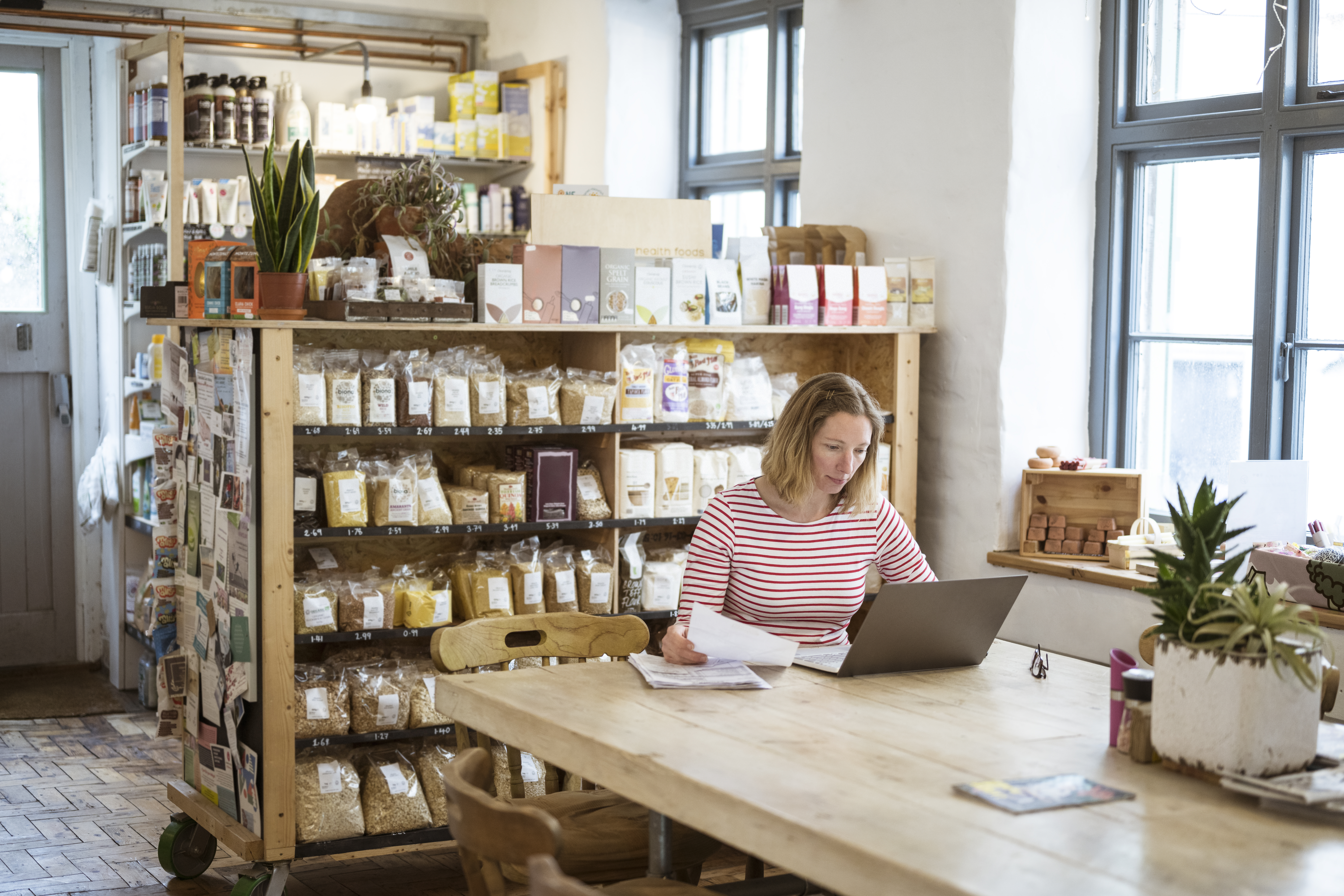Woman in early 40s using laptop at a communal table in a health food shop and cafe
