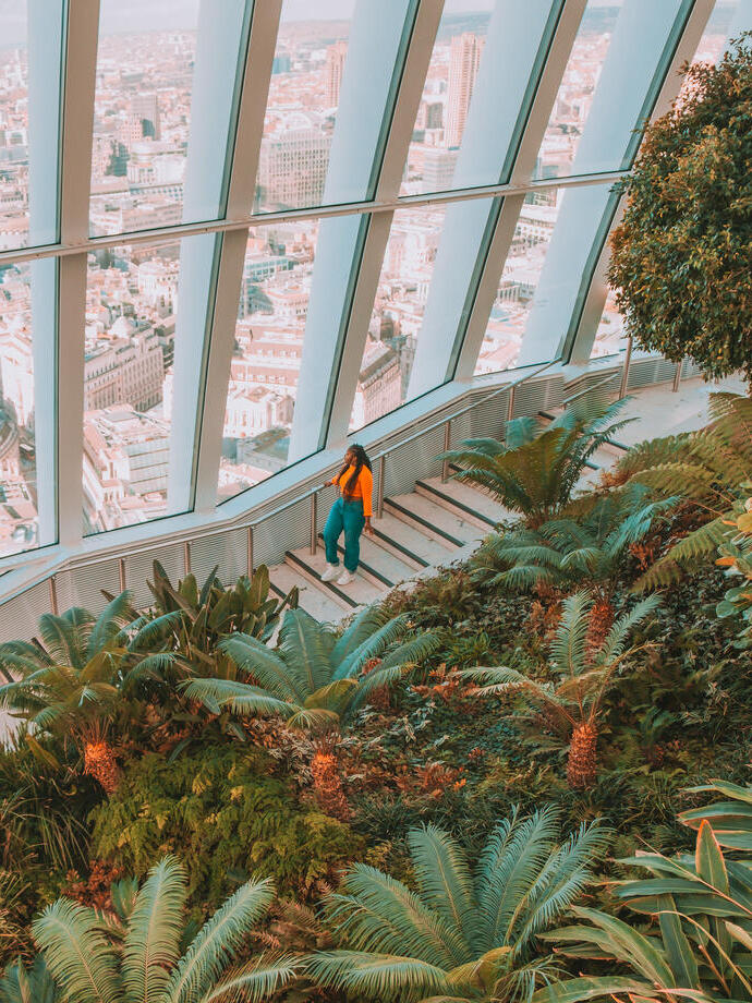 Woman stood surrounded by tropical plants looking out over the London skyline high up in a skyscraper.