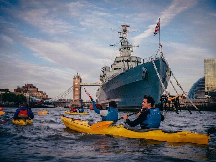 People in Kayaks on a river past iconic navy ships and iconic city landmarks.