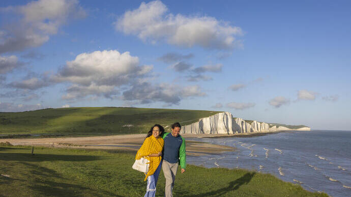 A couple walk together on a headland with white chalk cliffs in the background.