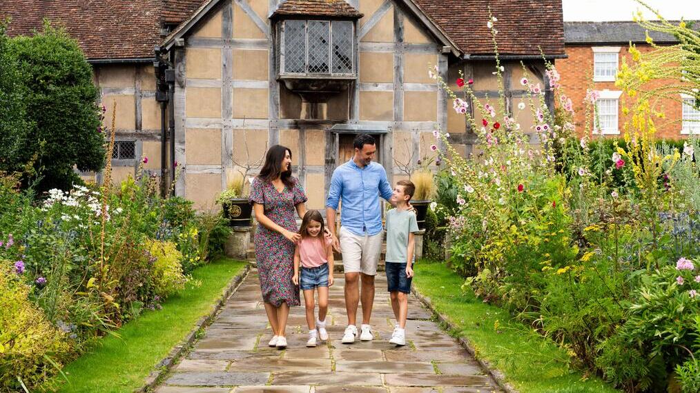 Eine Familie geht durch einen Garten vor einem historischen Haus mit schrägem Dach und blühendem Garten.