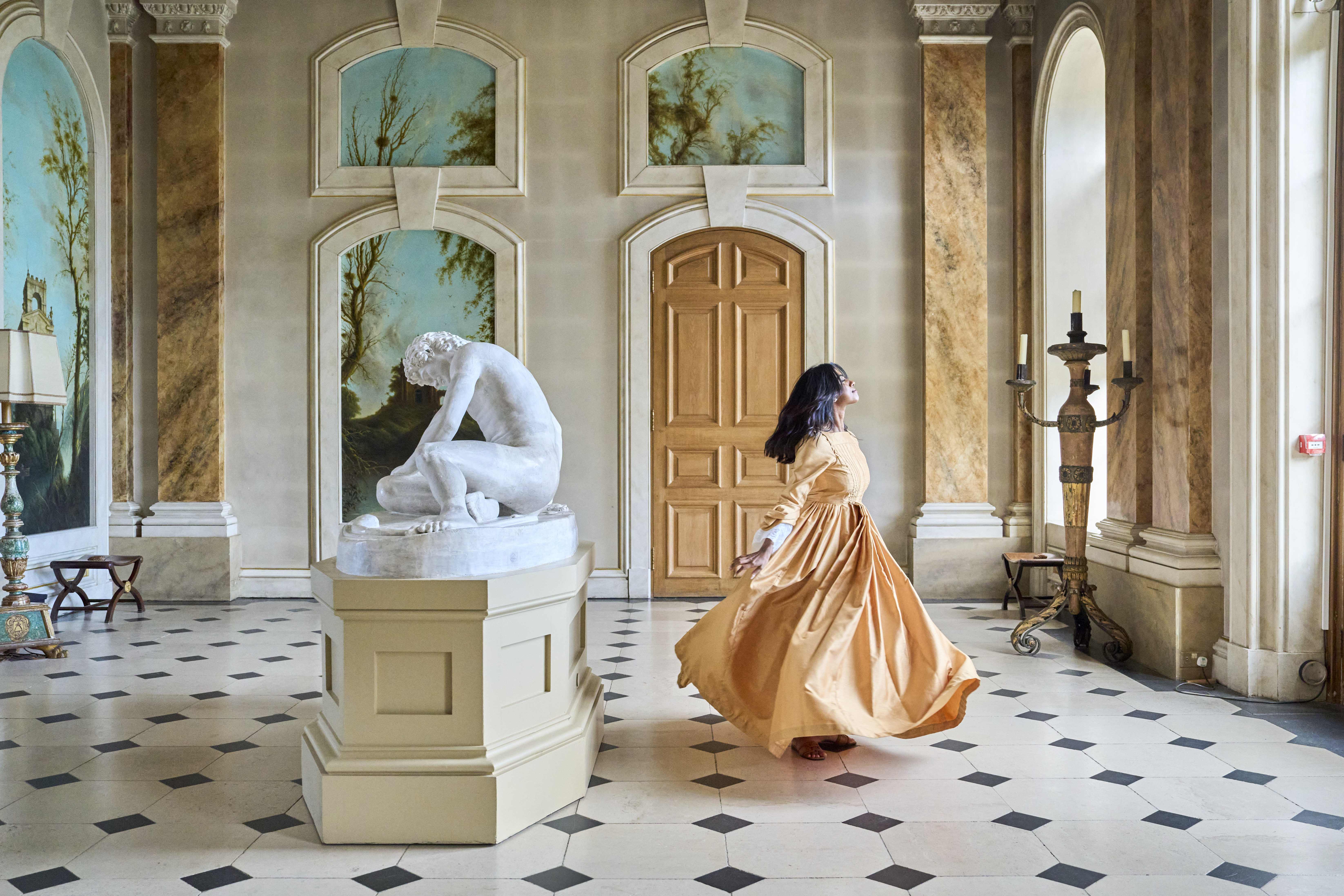 Elegant hall with classical statue, arched doors, large windows, and a person in a flowing period dress spinning gracefully on tiled floor.
