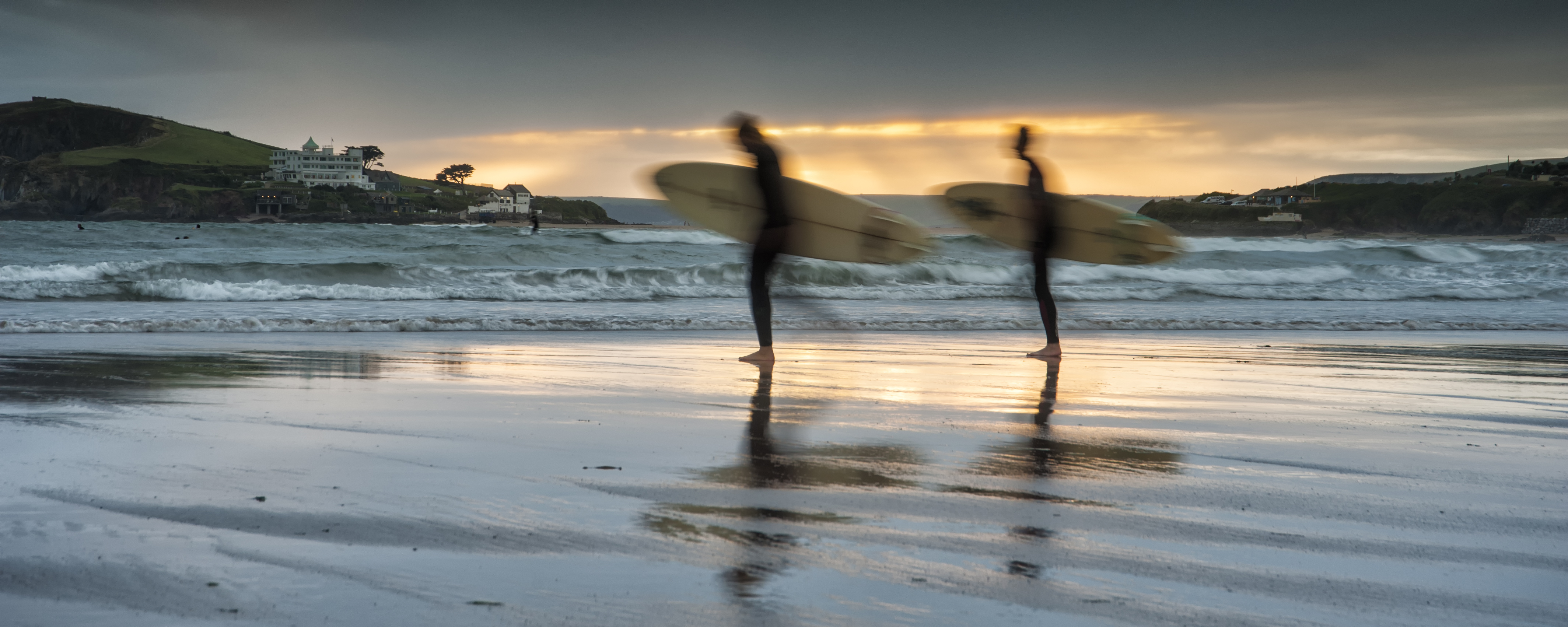 Two surfers with boards walk along a beach at sunset, waves and a coastal building visible in the background.