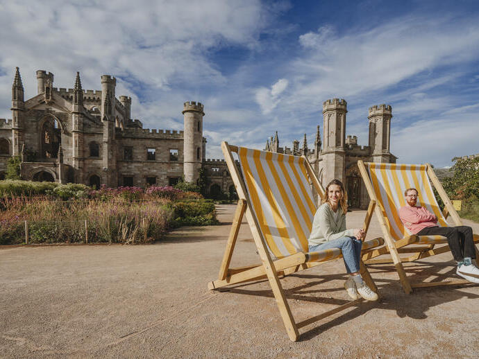 Deux personnes assises sur d’énormes chaises longues rayées jaune et blanc devant un grand château historique avec tours et jardins sous un ciel bleu.