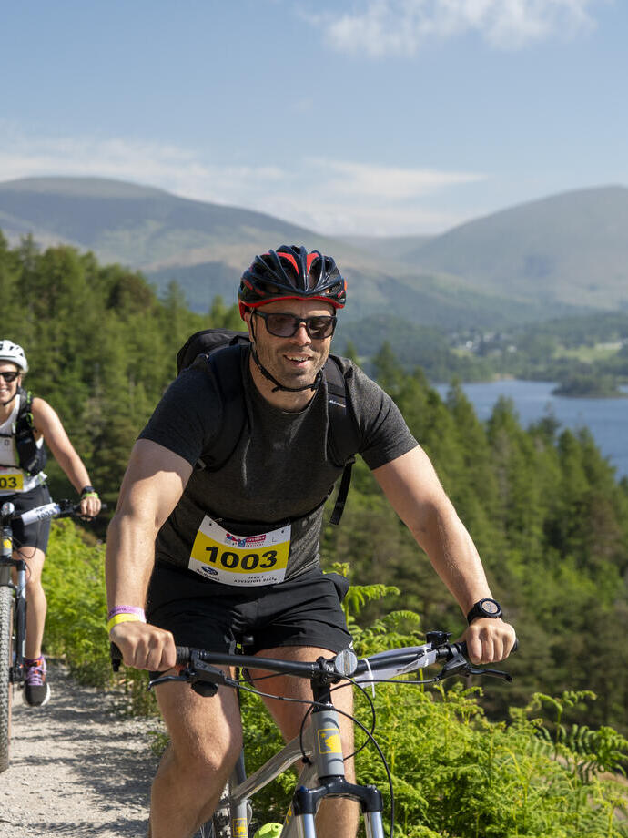 Two cyclists ride on a scenic trail beside a lake, surrounded by green hills and distant mountains under a blue sky.