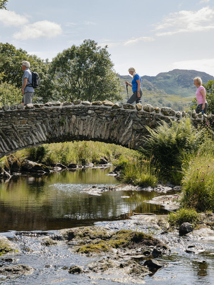 Un grupo de personas camina sobre un puente de piedra sobre un río, rodeado de paisaje verde y colinas bajo un cielo soleado.