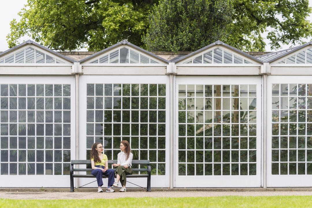 Two women sit on a bench in front of a glass pavilion