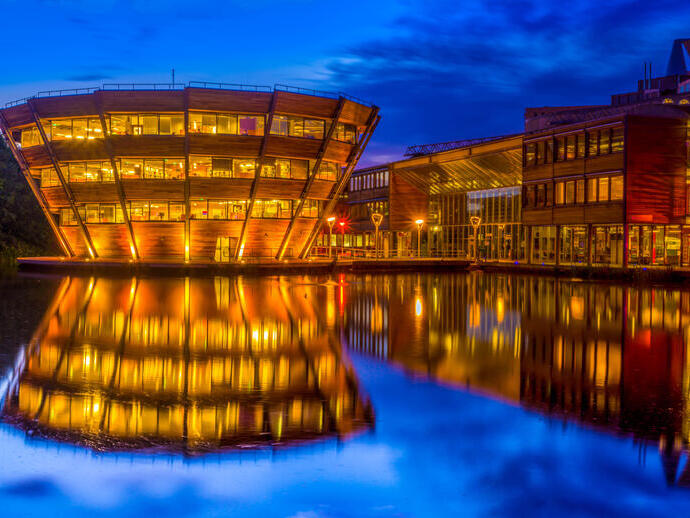 A modern, illuminated building with wooden panels is reflected in a calm body of water at night, against a deep blue sky.