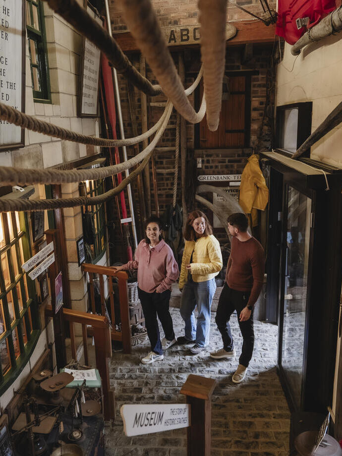 Three people inside a maritime museum with ropes, cobblestone floor, banners, and displays about local history and seafaring.