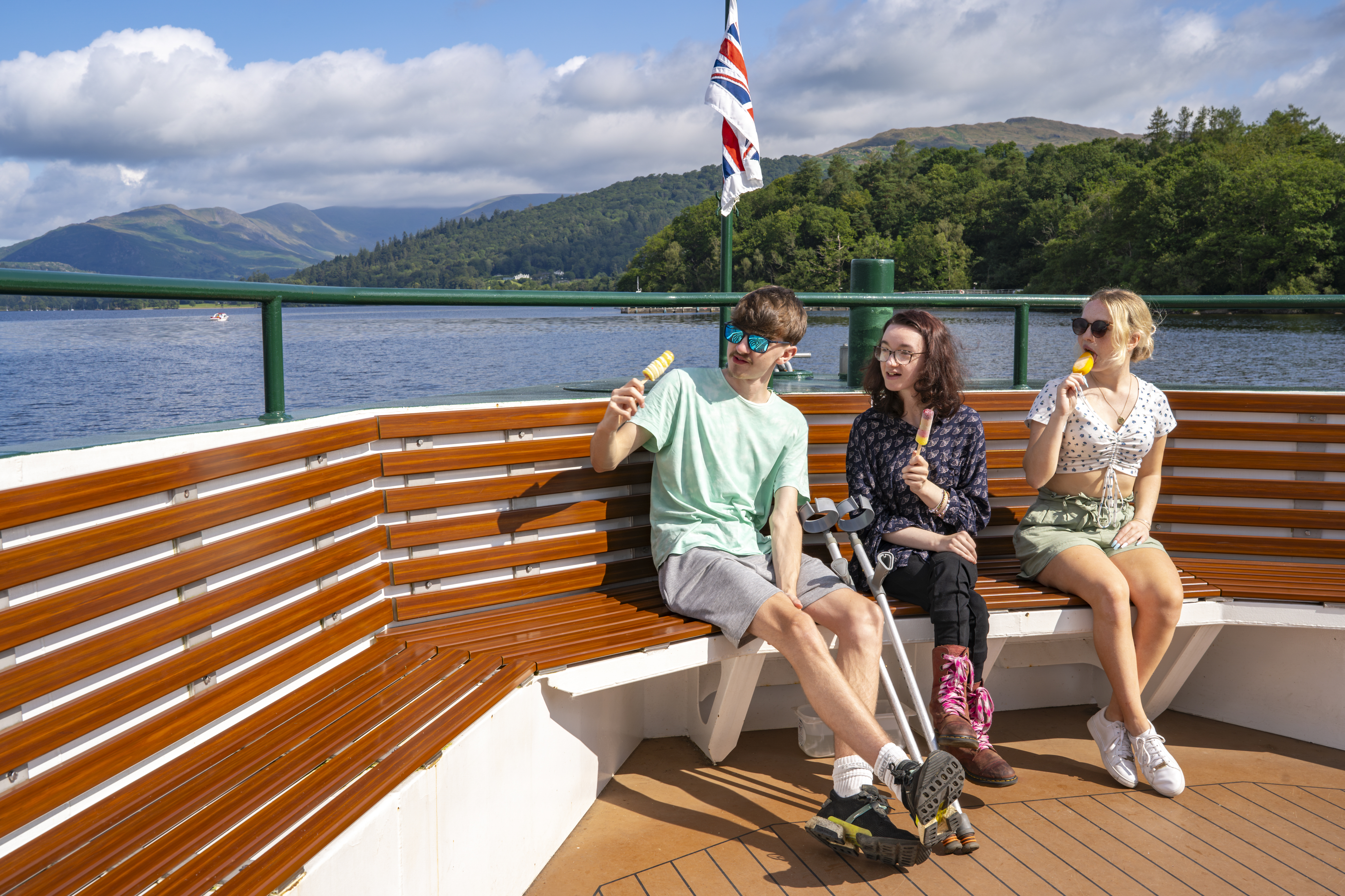 Three people sit at the end of a boat on a lake eating ice creams