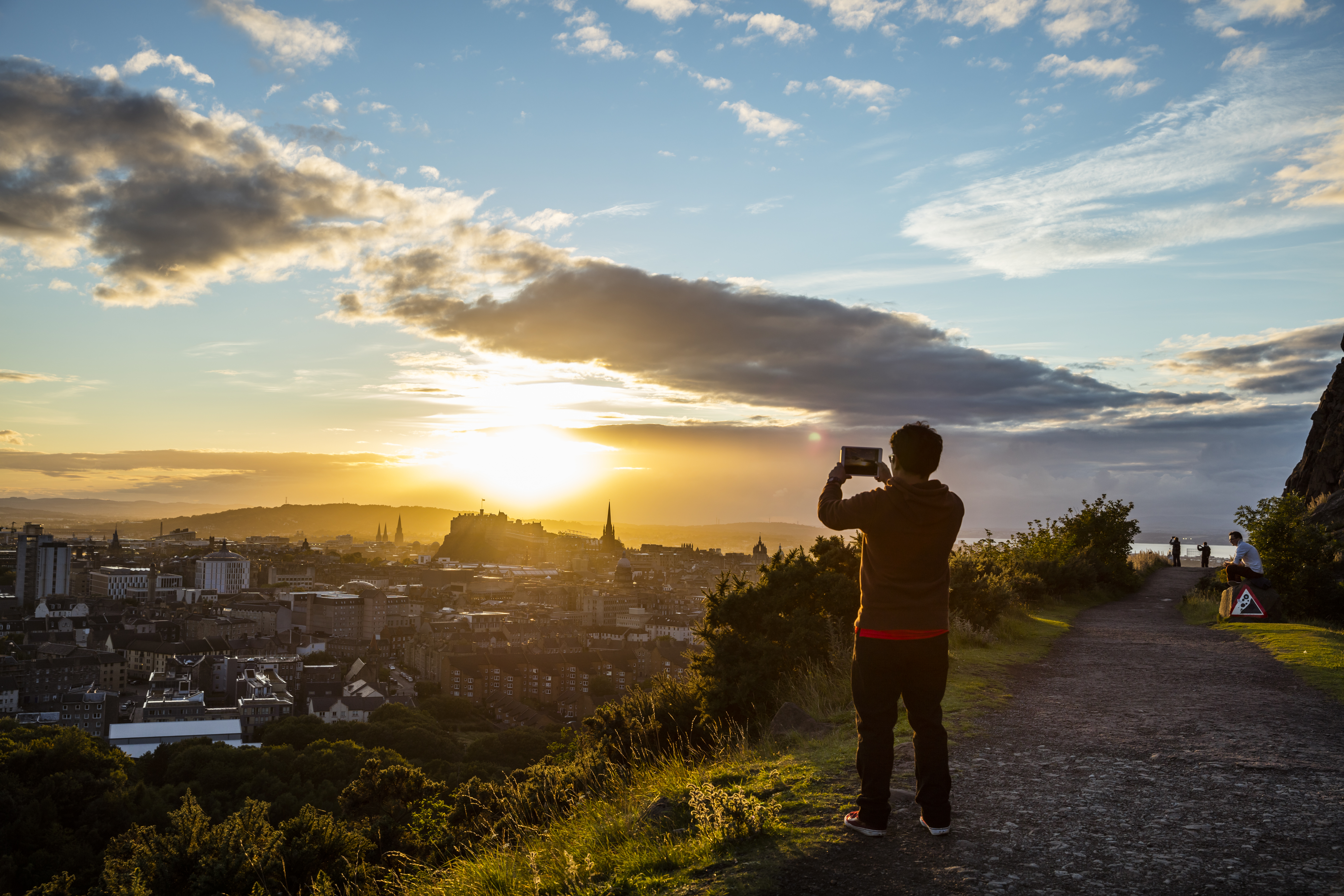 Jeune homme debout sur une falaise herbeuse prenant une photo du coucher de soleil