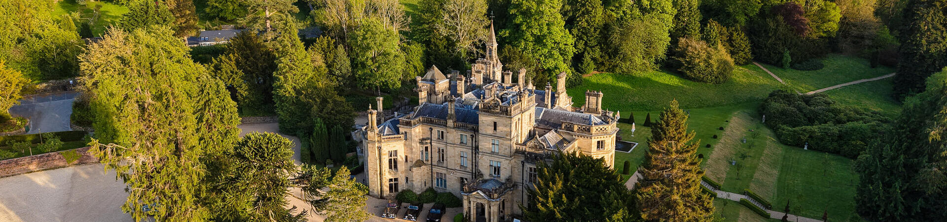 Aerial view of a grand historic manor surrounded by lush gardens, large trees, and rolling green hills under a blue sky.
