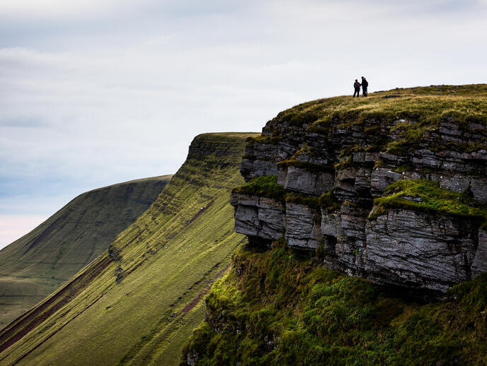 Two people stand atop a grassy cliff with dramatic rocky edges, overlooking rolling green hills under a cloudy sky.