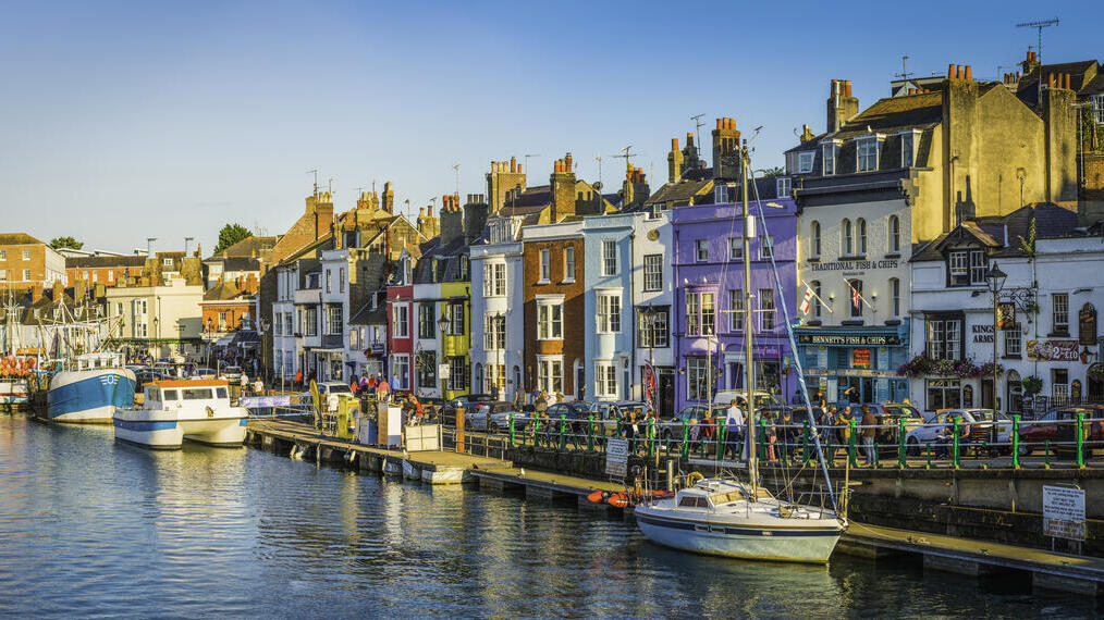 People walking up and down the water front of a busy seaside town lined with colourful terraced houses and boats anchored in the harbour