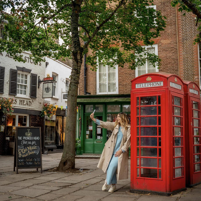 A woman leaning against a red telephone box and takes a selfie