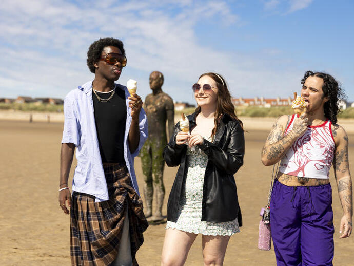 A group of friends walk on the beach with ice creams, a statue in the background