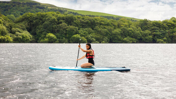 A woman kneels on a paddle board on a lake with countryside beyond