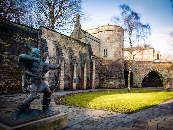 Statue of an archer in front of historic stone castle walls, green lawn, trees, and blue sky in the background.