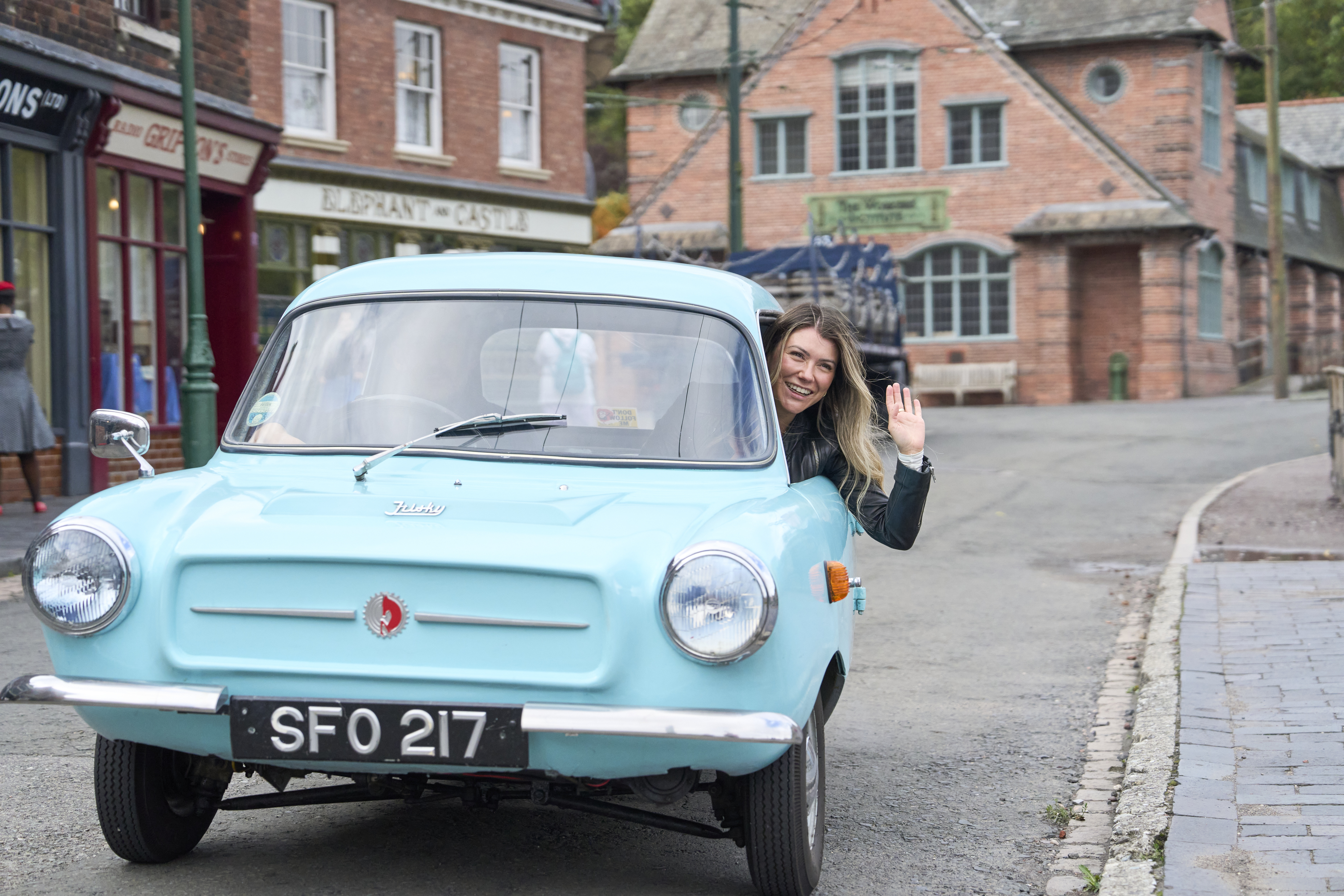 A woman waves from the window of a light blue vintage car on a quiet street with historic brick buildings and shops in the background.