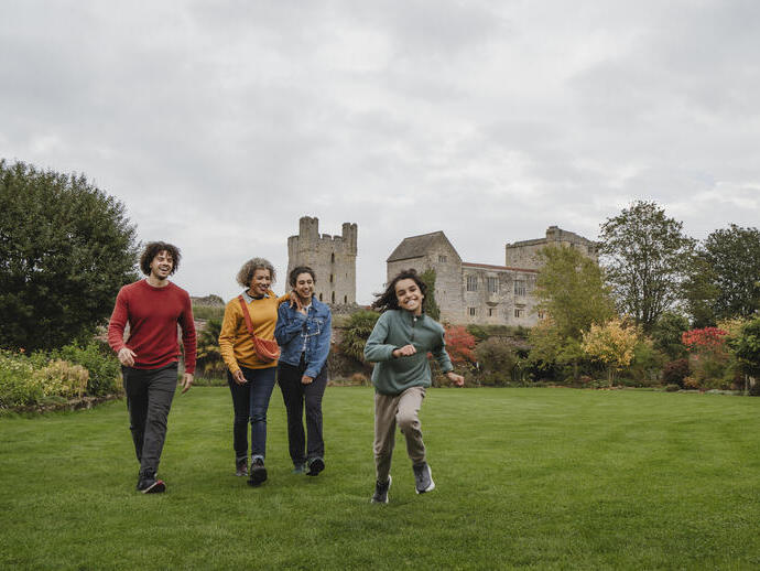 Four people walking on a large green lawn with an old stone castle and autumn trees in the background, under a cloudy sky.