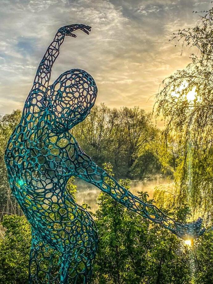 The sculpture Athena shown in long grasses beside the lake at a sculpture by the lakes exhibition in a botanical garden.