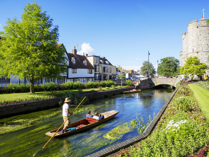 A man and a couple in a punt on a small river surrounded by gardens and houses