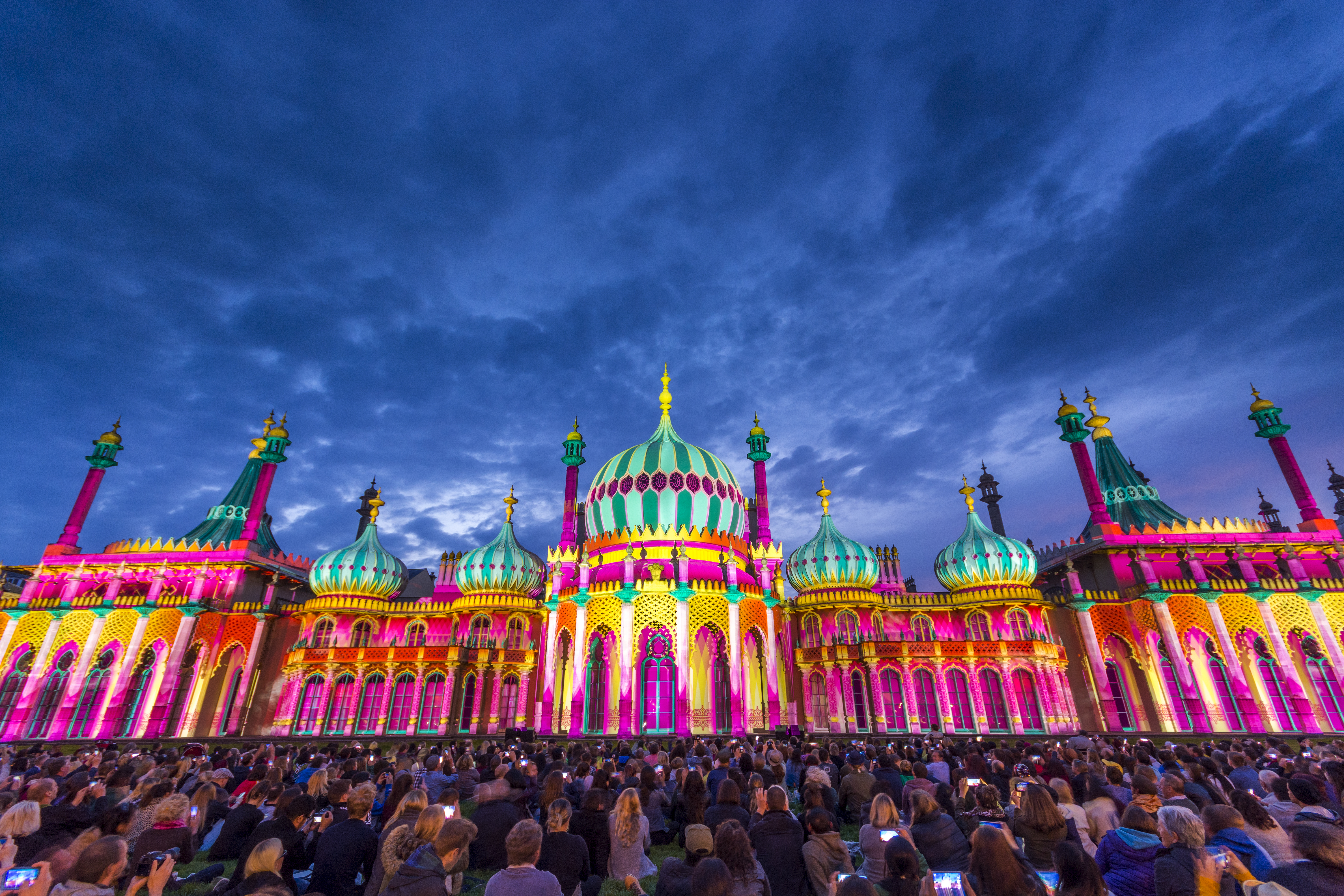 Crowd sitting on the ground in front of a large ornate building lit up with bright colours